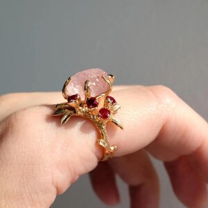 May include: A gold-toned ring featuring a large, light pink gemstone at the center, accented by smaller red gemstones. The ring's band and setting have a branch-like design, creating a nature-inspired aesthetic. The ring is displayed on a hand.