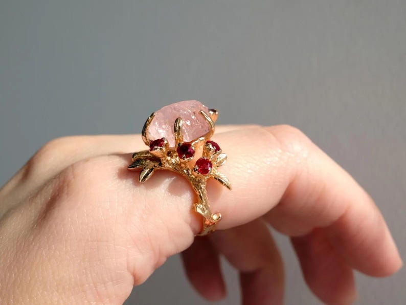 May include: A gold-toned ring featuring a large, light pink gemstone at the center, accented by smaller red gemstones. The ring's band and setting have a branch-like design, creating a nature-inspired aesthetic. The ring is displayed on a hand.