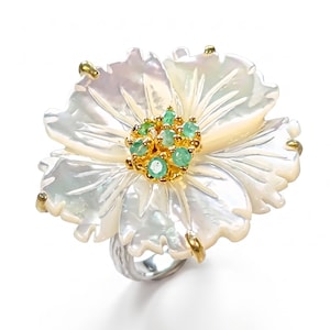 May include: A ring featuring a large, carved white mother-of-pearl flower with iridescent petals. The center of the flower is adorned with a cluster of small, round, green gemstones set in gold, with gold prongs securing the petals to a silver band.