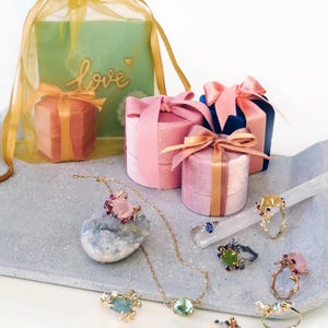 May include: Various pieces of jewellery, including rings and a necklace, are displayed on a grey tray. Several velvet ring boxes with satin bows are arranged around the jewellery. A gift bag with the word "love" is also present.