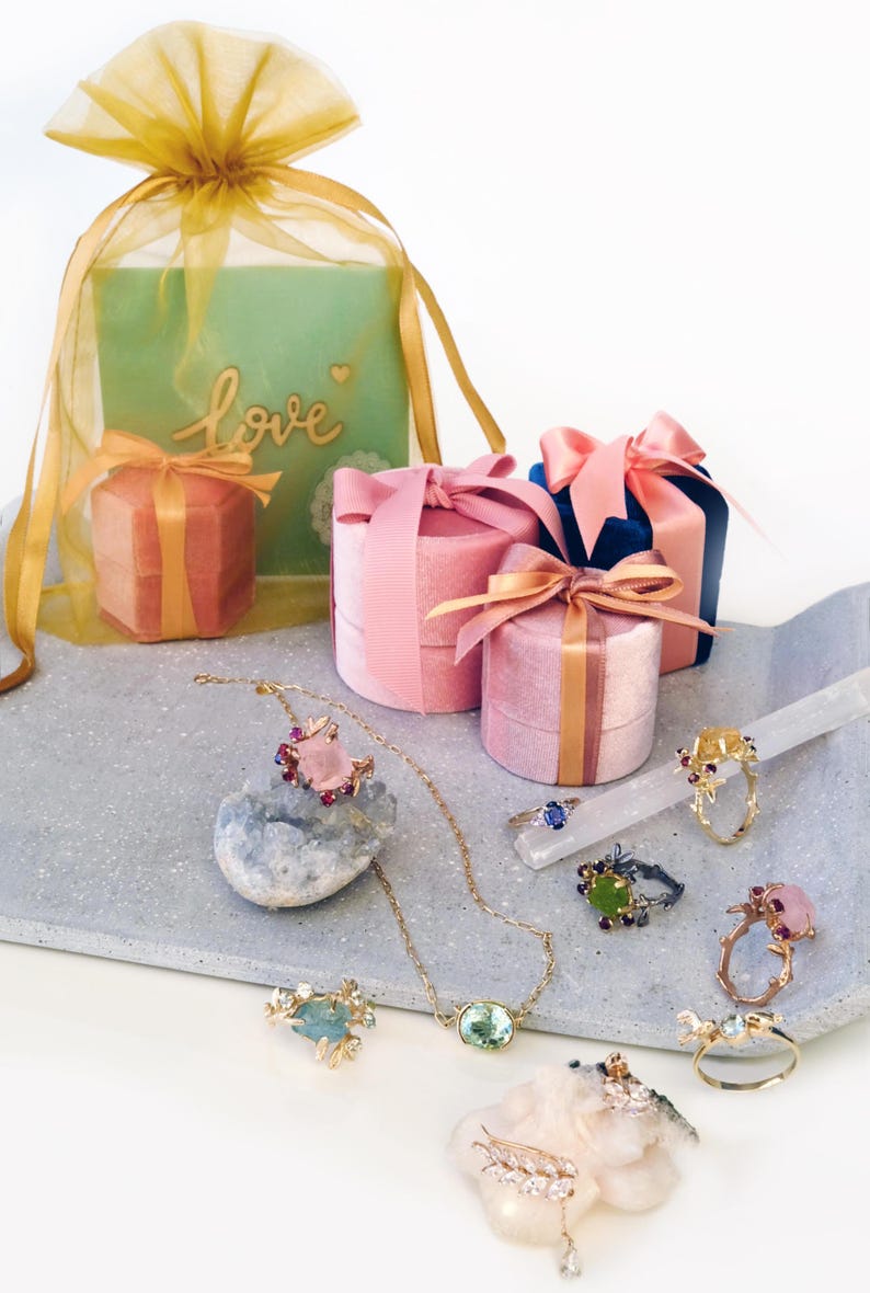 May include: Various pieces of jewellery, including rings and a necklace, are displayed on a grey tray. Several velvet ring boxes with satin bows are arranged around the jewellery. A gift bag with the word "love" is also present.