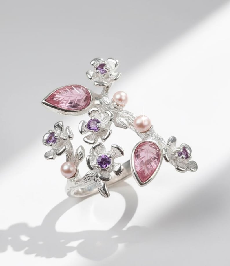 May include: A silver ring featuring a floral design with pink teardrop-shaped gemstones, purple accents, and small pink pearls. The ring has a detailed, nature-inspired aesthetic.