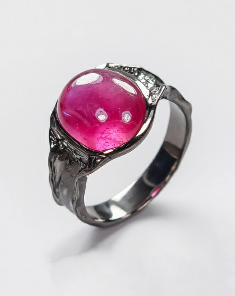 May include: A dark grey ring with a large, vivid pink gemstone. The oval-shaped gemstone is set in a textured, ornate metal band. The ring's design includes decorative elements around the gemstone, enhancing its unique aesthetic.