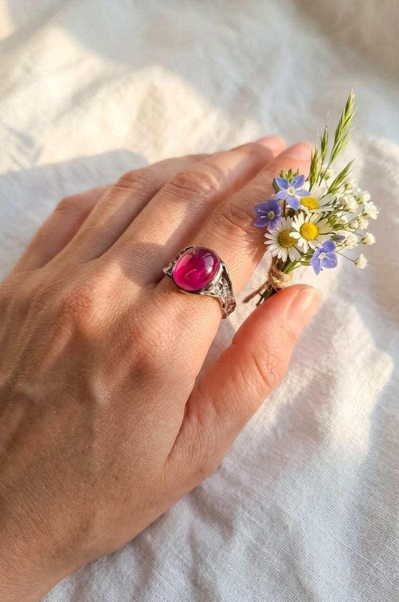 May include: A silver ring featuring a large, oval, magenta gemstone. The ring is worn on a hand, with a small posy of wildflowers held close. The ring's setting has an intricate, filigree pattern.