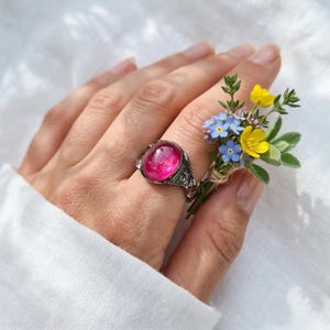 May include: A silver ring featuring a large, oval, hot pink gemstone. The ring has an ornate, detailed design. A small bouquet of yellow and blue wildflowers is held nearby.