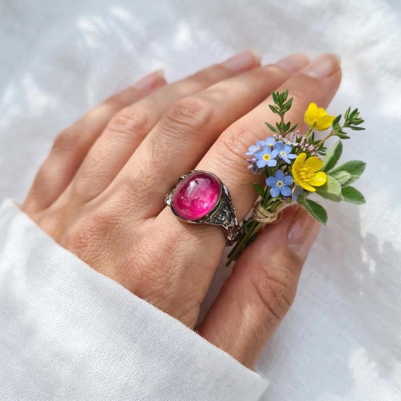 May include: A silver ring featuring a large, oval, hot pink gemstone. The ring has an ornate, detailed design. A small bouquet of yellow and blue wildflowers is held nearby.