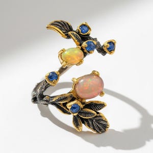 Könnte beinhalten: Silberfarbener Ring mit floralem Design, mit zwei ovalen Opalen und mehreren kleinen blauen Edelsteinen. Der Ring hat goldfarbene Akzente und schwarze Details, was eine von der Natur inspirierte Ästhetik schafft.