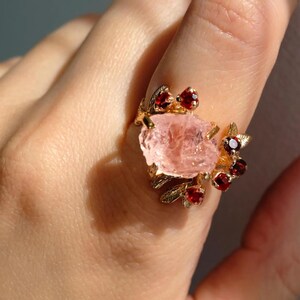 May include: A gold-toned ring featuring a large, rough-cut pink gemstone as the centerpiece. The ring is adorned with smaller, round, red gemstones and delicate leaf-like details.