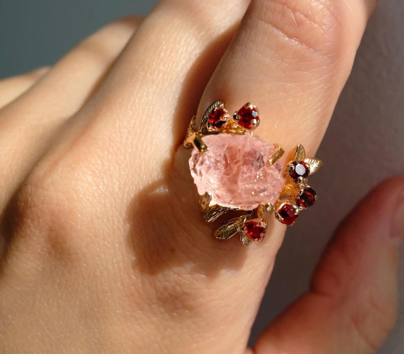 May include: A gold-toned ring featuring a large, rough-cut pink gemstone as the centerpiece. The ring is adorned with smaller, round, red gemstones and delicate leaf-like details.