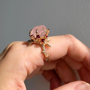 May include: A gold-colored ring featuring a large, light pink gemstone at the center, surrounded by smaller red gemstones. The ring has a branch-like design with intricate details, set against a neutral background.