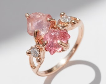 Può includere: Un anello in oro rosa con tre pietre preziose rosa e due pietre trasparenti più piccole. L'anello presenta una grande pietra rosa rettangolare e una pietra rosa più piccola e appuntita, con una pietra trasparente su entrambi i lati. L'anello è su uno sfondo bianco.