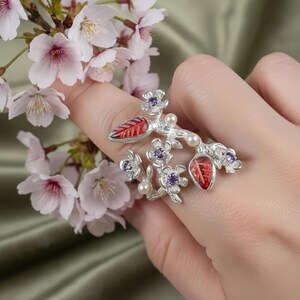 May include: A silver ring with a floral design featuring red enamel leaves, purple gemstones, and small pearls. The ring is set against a backdrop of pink cherry blossoms, creating a nature-inspired aesthetic.