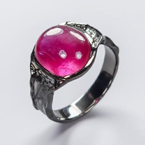 May include: A dark gray ring with a large, round, magenta-colored stone. The ring has a textured band and decorative accents around the stone. The stone appears to be smooth and polished, reflecting light.