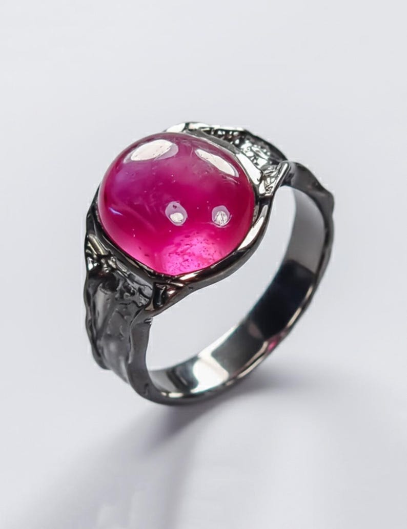 May include: A dark grey ring featuring a large, round, magenta gemstone. The ring has a textured band and decorative detailing around the stone. The polished stone reflects light, creating a striking contrast.