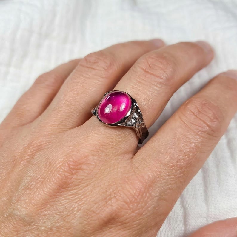 May include: A silver ring with a large, oval, bright pink gemstone. The ring has a detailed, ornate band with a dark finish. The gemstone is the focal point, catching the light and adding a vibrant colour to the piece.