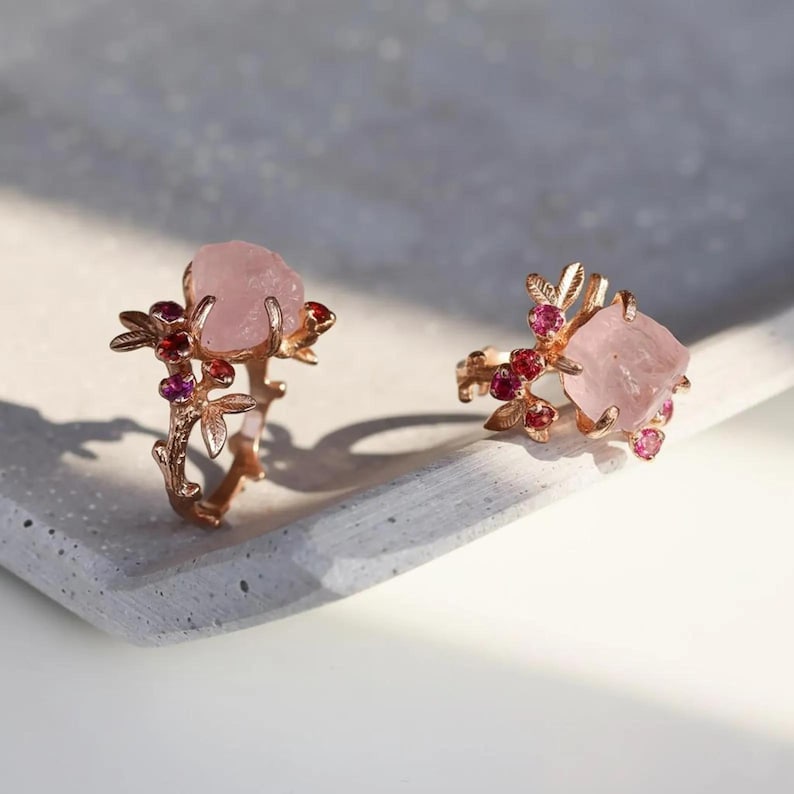 May include: Two rose gold rings with large, light pink gemstones. The rings feature a floral design with small red and pink accent stones. The rings are displayed on a light gray surface, showcasing the intricate details and colors of the jewelry.