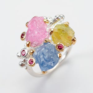 Peut inclure: Une bague en argent ornée de trois grosses pierres brutes non taillées de couleur rose, jaune et bleue. Le sertissage est agrémenté de petites pierres rouges rondes et facettées fixées par des griffes dorées sur une structure en argent texturé.