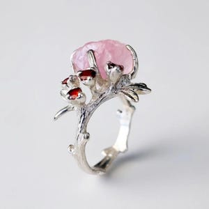 May include: A silver ring featuring a large, light pink gemstone at the top, resembling a flower. The ring's band and setting are designed to look like a tree branch, with small red gemstones accenting the design. The ring has a nature-inspired theme.