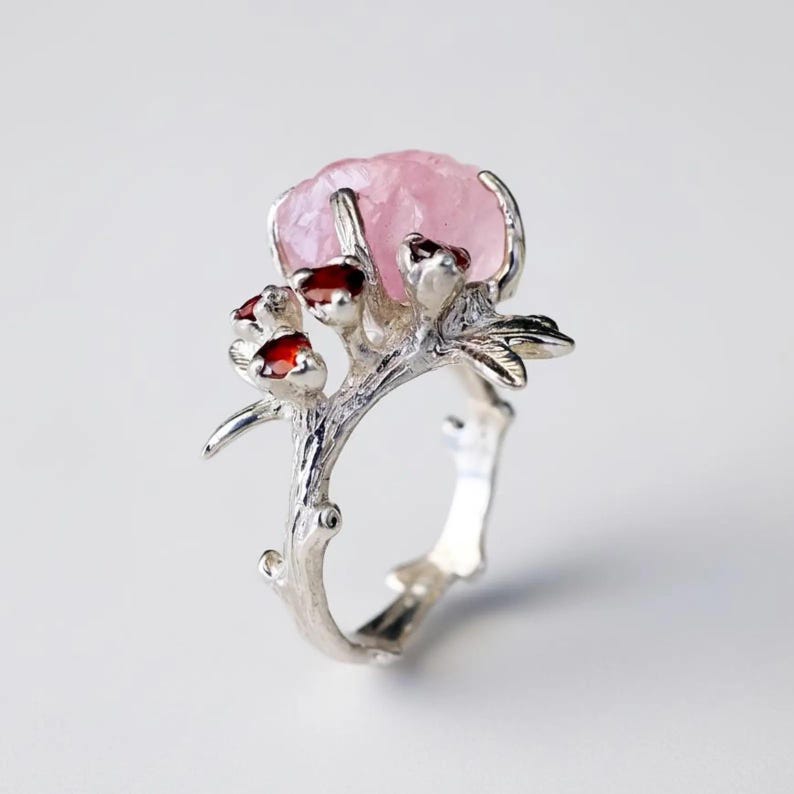 May include: A silver ring featuring a large, light pink gemstone at the top, resembling a flower. The ring's band and setting are designed to look like a tree branch, with small red gemstones accenting the design. The ring has a nature-inspired theme.