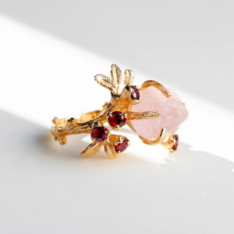 May include: A gold-colored ring featuring a branch design with leaves and red gemstone accents. A large, light pink, irregularly shaped gemstone is the focal point. The ring is set against a white background, highlighting its intricate details.