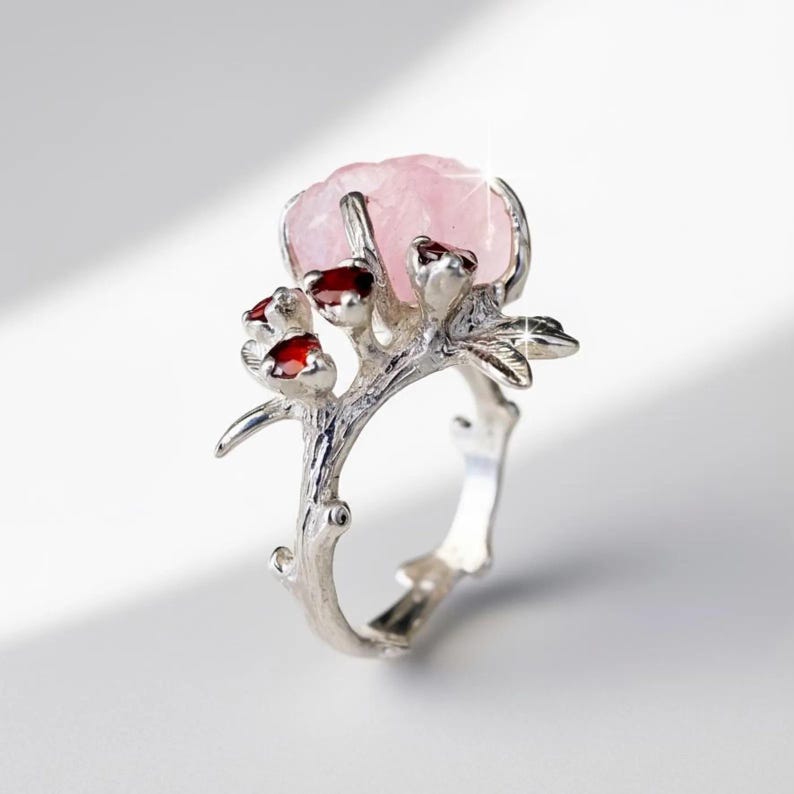 May include: A silver ring featuring a large, light pink gemstone at the center, resembling a flower. The ring's band and setting are designed to look like a branch with small red accents.