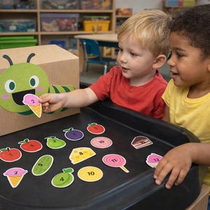 Peut inclure: Deux enfants interagissent avec une boîte en carton décorée d'une chenille verte. La boîte a une découpe en forme de bouche pour nourrir des cartes numérotées en forme d'aliments, dont une glace. Des cartes colorées de fruits et d'aliments sont éparpillées sur une surface noire.