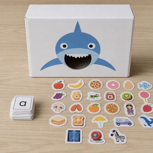 Peut inclure: Une bo&icirc;te blanche avec une illustration de requin bleu, et une pile de cartes alphab&eacute;tiques. Des autocollants assortis d'aliments et d'objets sont dispers&eacute;s autour de la bo&icirc;te, dont une pizza, un z&egrave;bre et une fraise. Jouet &eacute;ducatif.