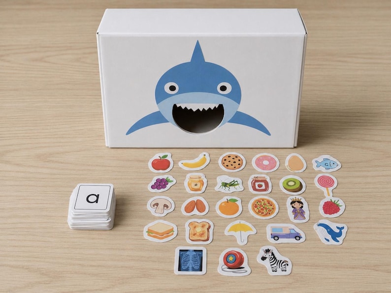 Peut inclure: Une bo&icirc;te blanche avec une illustration de requin bleu, et une pile de cartes alphab&eacute;tiques. Des autocollants assortis d'aliments et d'objets sont dispers&eacute;s autour de la bo&icirc;te, dont une pizza, un z&egrave;bre et une fraise. Jouet &eacute;ducatif.