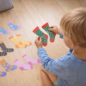 Może przedstawiać: Dziecko trzyma dwie zielone skarpetki w białe grochy i czerwone mankiety. Kilka par kolorowych skarpetek jest rozrzuconych na drewnianej podłodze, z różnymi wzorami i wzorami, w tym paski, serca i uśmiechnięte buzie.