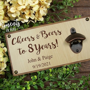 May include: Wooden sign with a bottle opener attached. The sign is light brown with dark brown text that reads "Cheers & Beers to 8 Years!"  John & Paige 9/19/2021.