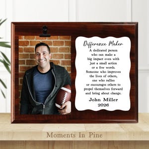 May include: A wooden picture frame with a photo of a man holding a football and a printed message. The frame has a dark brown finish and a clip at the top. The text on the right side reads "Difference Maker".