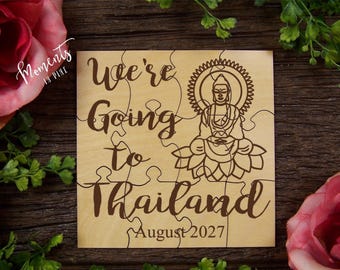 Può includere: Un puzzle quadrato in legno con testo inciso al laser in marrone scuro che recita We're Going to Thailand August 2027. Il design include un'illustrazione dettagliata a linee di una figura di Buddha seduta su una base a fiore di loto.