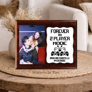 May include: A rectangular dark wood plaque featuring a photo clip on the left and a white panel on the right. The white panel displays the text FOREVER IN 2 PLAYER MODE, two game controllers with a heart, and the names BRAD AND CHANTELLE 04.10.2026.