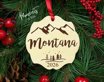 Può includere: Un ornamento in legno chiaro con bordo smerlato, caratterizzato da una grafica di montagna marrone scuro, il testo "Montana", una piccola illustrazione di una foresta e l'anno "2026". L'ornamento è appeso a un nastro rosso.