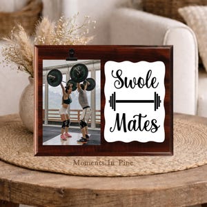 May include: A rectangular dark wood photo frame with a black metal clip holding a photograph of two people lifting barbells. To the right, a white scalloped sign features the black text "Swole Mates" above and below a black barbell icon.