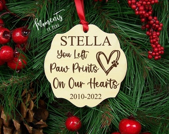 May include: A light-colored wooden ornament with a scalloped edge, hanging from a red ribbon. It features dark brown engraved text reading STELLA, You Left Paw Prints On Our Hearts, and 2010-2022, accompanied by a heart graphic with a paw print.