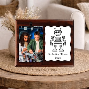May include: A rectangular dark wood plaque featuring two side-by-side sections. The left side displays a photograph of two people working on a robot. The right side is a white scalloped panel with a black robot illustration and the text Robotics Team 2026.