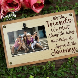 May include: Wooden picture frame with a light brown background and a photo of a group of friends sitting on steps. The frame has a clip at the top and the text "It's the Friends We Meet Along the Way That Helps Us Appreciate the Journey." is printed on the frame.