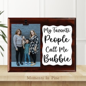 May include: A dark brown wooden picture frame with a photo of a family and the text "My Favorite People Call Me Bubbie." The frame has a clip to hold the photo and is on a wooden surface.