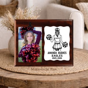 May include: A rectangular dark wood plaque featuring a clip holding a photograph of a cheerleader with red pom-poms. Beside it, a white scalloped insert displays a black graphic of a cheerleader and the text AMANDA HUGHES EAGLES 2025-2026.