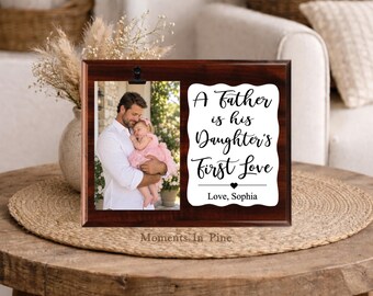 Può includere: Una cornice rettangolare in legno scuro con una clip in metallo nero che sostiene la fotografia di un uomo che tiene in braccio un bambino. A destra, una placca bianca smerlata riporta il testo nero: A Father is his Daughter's First Love, Love, Sophia.