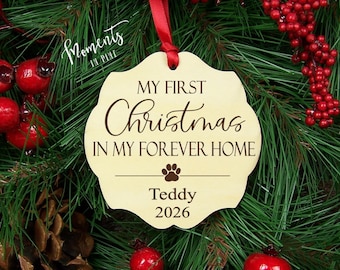 Puede incluir: Un adorno de madera con bordes festoneados y una cinta roja, grabado con el texto MY FIRST Christmas IN MY FOREVER HOME, un icono de huella, el nombre Teddy y el año 2026. El adorno está centrado sobre un fondo de agujas de pino.