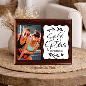 May include: A rectangular dark wood photo frame featuring a black metal clip to hold a photograph of two people. To the right, a white scalloped plaque displays the black text Sole Sisters, the names Amy & Shawna, and decorative leaf motifs.
