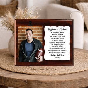 May include: A rectangular dark wood plaque featuring a black metal clip holding a photograph of a person holding a football against a brick wall. Beside the photo is a white scalloped card with the text Difference Maker, John Miller, 2026.