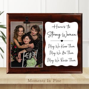 May include: A dark brown wooden picture frame with a photo of three people and a white sign with black text that reads "Here's to Strong Women. May We Know Them, May We Be Them, May We Raise Them."
