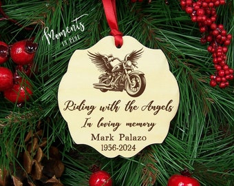 May include: A light-colored wooden ornament with a scalloped edge, featuring a dark brown engraved graphic of a motorcycle with wings. The text reads: Riding with the Angels, In loving memory, Mark Palazo, 1956-2024. A red ribbon is attached at the top.
