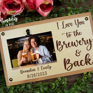 May include: A wooden picture frame with a clip to hold a photo. The frame has a light brown background with the text "I Love You to the Brewery & Back" and the words "Moments in Pine" at the top. The frame also has a date and names at the bottom. The photo shows a couple holding beers in a bar.