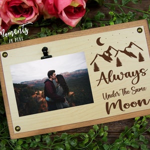 May include: A wooden photo frame with a clip to hold a photo. The frame has a brown stain and a white background. The frame has the text "Always Under The Same Moon" and a mountain and moon design.