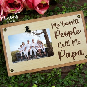 May include: Wooden picture frame with a clip at the top. The frame has a light brown background with a darker brown border. The frame has a photo of a family of five sitting on a bench. The text "My Favorite People Call Me Papa" is written in brown on the frame.