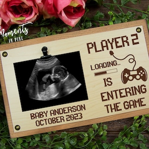 May include: A wooden sign with a photo frame and the text "Player 2 is entering the game." The frame contains an ultrasound image. The sign also includes the text "Baby Anderson October 2023".